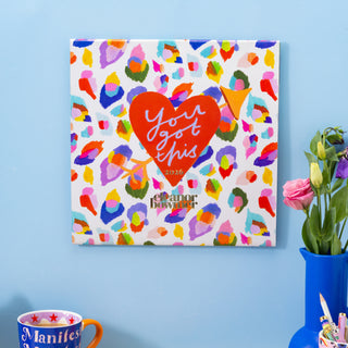 Colorful calendar by Eleanor Bowmer with heart design and text on a blue wall, next to a mug and vase with flowers.