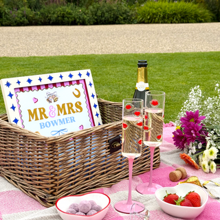 Picnic setup with champagne, glasses, and personalized sign on a grassy area.
